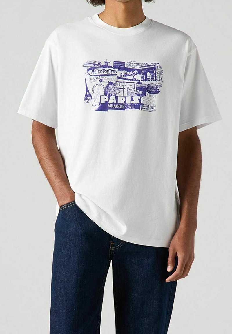 Personne portant un T-shirt blanc avec des imprimés graphiques bleus représentant des monuments et panneaux de Paris, associé à un jean bleu foncé.