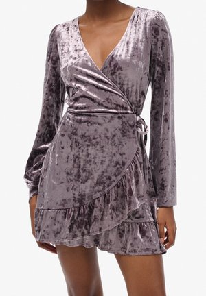 Femme portant une robe portefeuille en velours froissé violet à manches longues avec un ourlet volanté et une fermeture nouée sur le côté.