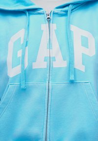 Ανοιχτό γαλάζιο φούτερ με φερμουάρ, με λευκά γράμματα "GAP", κορδόνια και μπροστινή τσέπη τύπου καγκουρό.