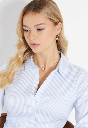 Gouden oorbellen in de vorm van bloemen en een bijpassende ketting gedragen met een lichtblauwe gestreepte blouse. Glad stof, delicate kettingdetails.
