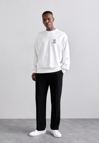 Weißes Sweatshirt mit grünem Grafikdesign auf der Brust, kombiniert mit schwarzen Hosen und weißen Sneakers. Glattes Material und lässiger Schnitt.