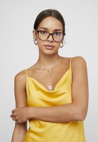 Camisón de satén amarillo con escote drapeado, collares de plata en capas y gafas de carey con lentes de tintado azul.
