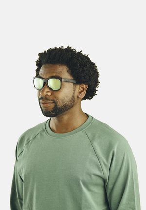 Zonnebril met donkere monturen en groene reflecterende lenzen. Draagt een persoon in een lichtgroen sweatshirt met een ronde hals en lange mouwen.