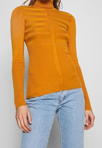 Pull à col roulé moutarde ajusté avec des motifs en tricot côtelé et rayé, associé à un jean taille haute bleu clair porté par une personne debout.