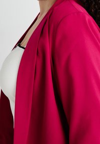 Tight vit topp under en lös, livlig rosa ytterjacka. Tyget verkar slätt med en lätt glans och subtila sömnadsdetaljer.