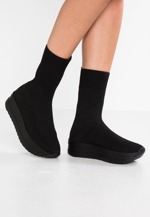 Person iført sorte strømpe-lignende platformsneakers med tekstureret strikoverdel, tykke sorte såler, stående på et hvidt gulv.