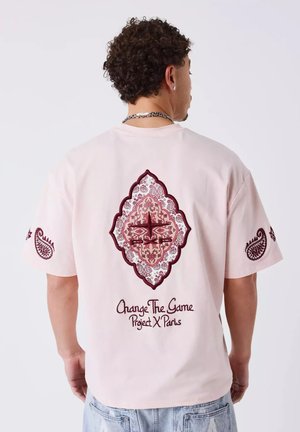 Man met krullend haar die een lichtroze T-shirt draagt met wijnrode paisleyprint en de tekst "Change The Game Project X Paris" op de rug.