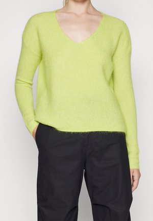 Persona vistiendo un suéter de cuello en V color verde lima con mangas largas y pantalones negros, con las manos parcialmente en los bolsillos, sobre un fondo liso.