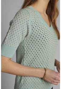 Pull-over en maille vert menthe clair avec un col en V, présentant des trous texturés sur toute la surface et des manches courtes. Agrémenté d'un ourlet et de poignets côtelés.