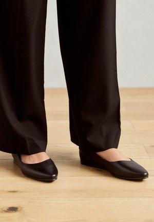 Black leather ballet flats with a rounded toe and minimalistic design, paired with loose black pants on a wooden floor.
