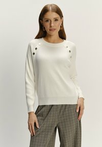 Sweter dzianinowy w kolorze kremowym z żeberkową teksturą, z metalowymi guzikami na ramieniu. Połączony z spodniami w kratę dla kontrastu.