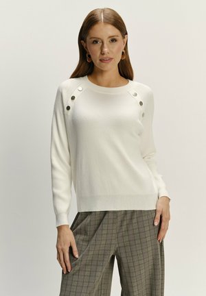 Sweter dzianinowy w kolorze kremowym z żeberkową teksturą, z metalowymi guzikami na ramieniu. Połączony z spodniami w kratę dla kontrastu.