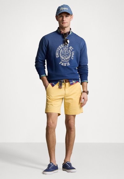 Sweatshirt azul com gráfico branco, sobre uma camisa xadrez. Calções amarelos e sapatilhas de tela azuis completam o visual. O boné combina com o sweatshirt.