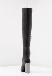 Bota alta de cuero negro con textura suave, punta cuadrada y tacón cuadrado de plata. Presenta un cierre lateral y detalles de costura en la parte posterior.