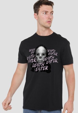 Mann trägt schwarzes T-Shirt mit silberner Totenkopfmaske und wiederholtem Text "Death Eater", blickt seitlich vor einfarbigem Hintergrund.