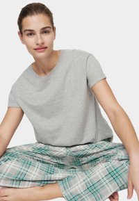 Joven con el cabello recogido, vestida con una camiseta gris y pantalones a cuadros verdes, sentada con las piernas cruzadas frente a un fondo simple.