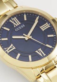 Guld metalurisk ur med en rund marineblå urskive, romerske talmarkører og guldhænder. Glat armbåndsdesign med poleret finish.