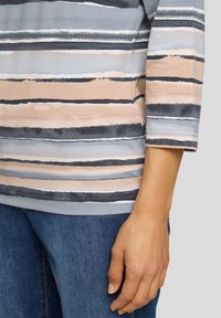 Nahaufnahme einer Person, die Blue Jeans und ein Oberteil mit Dreiviertelärmeln in horizontalen grauen, beigen und schwarzen Streifen trägt, mit entspanntem Arm an der Seite.