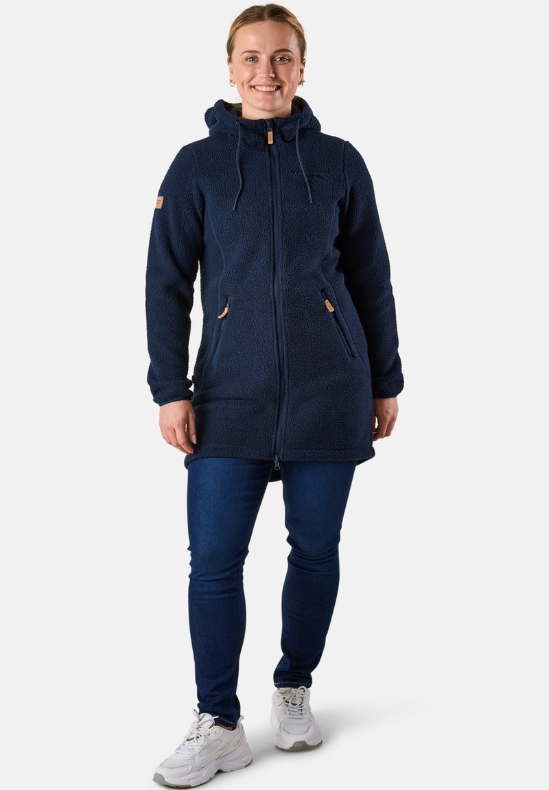 Kvinde står iført en marineblå fleecehættejakke, mørkeblå jeans og hvide sneakers, mens hun smiler foran en ensfarvet baggrund.