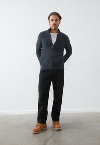 Pull en maille zippé gris foncé avec une finition texturée, porté par-dessus une chemise blanche, associé à un pantalon noir et des chaussures à lacets marron.