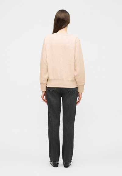 Kvinne med langt brunt hår iført en beige genser og svarte jeans med rette ben, stående med ryggen mot på en ensfarget hvit bakgrunn.