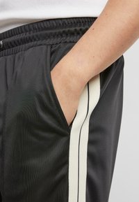 Pantalon de sport noir avec des rayures blanches sur les côtés, en tissu léger, doté d'une taille élastique et d'une poche latérale.