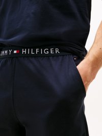 Mann in dunkelblauen Tommy Hilfiger-Hosen mit schwarzem Bund und Markennamen, Hand in der Tasche, vor einem hellen Hintergrund.