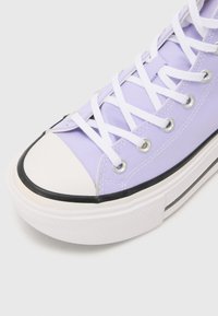 Basket haute en toile lilas avec des lacets blancs, des œillets argentés et une semelle en caoutchouc noire. Pointe arrondie avec un capuchon blanc, surface texturée.