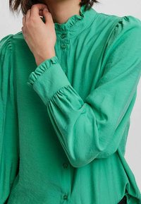 Grüne Bluse mit langen Ärmeln aus leichtem Stoff. Sie verfügt über einen Rüschenausschnitt und eingefasste Manschetten sowie ein Knopfverschluss vorne.