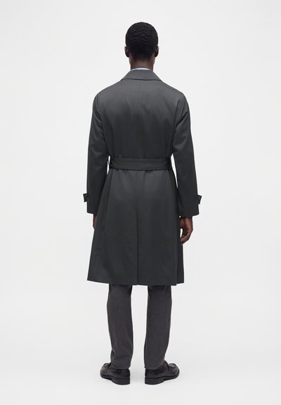 Homme debout de dos, portant un trench-coat gris foncé ceinturé, un pantalon gris et des chaussures habillées noires sur un fond uni.