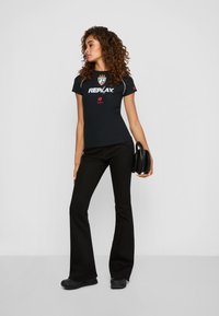 Vrouw met krullend haar die een zwart Replay logo T-shirt, zwarte wijde broek, sneakers draagt en een zwarte handtas vasthoudt, terwijl ze zijwaarts poseert.