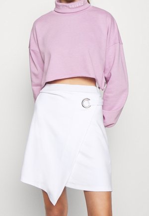 Femme portant un sweat-shirt court lavande avec "Missguided" sur le col et une jupe portefeuille asymétrique blanche avec un détail anneau argenté.