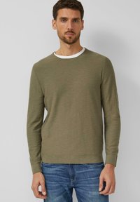 Mężczyzna w oliwkowozielonym swetrze z długim rękawem, nałożonym na białą koszulę, w niebieskich dżinsach, stojący na tle jednolitego jasnoszarego tła.