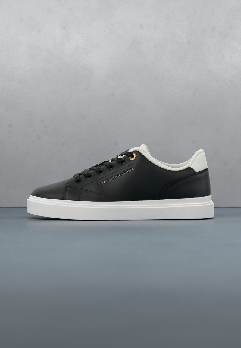Baskets basses noires avec semelle blanche et col de talon, lacets noirs et logo Tommy Hilfiger doré sur le côté sur fond gris.