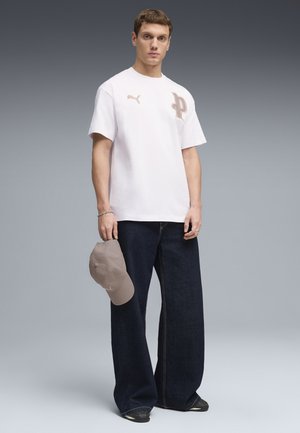 Junger Mann steht und hält eine beige Kappe, trägt ein weißes Puma T-Shirt mit Logo und lockere dunkelblaue Jeans vor einem grauen Hintergrund.