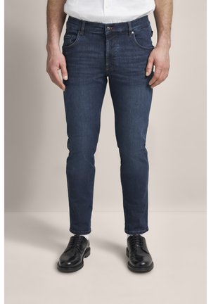 Dunkelblaue Denim-Jeans mit Slim-Fit, ausgestattet mit vorderen Taschen und subtilen Nähdetails, kombiniert mit polierten schwarzen Schuhen.