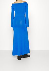 Långärmad klänning med bar axel i starkblå knittrad tyg, åtsittande design med synliga sömmar, kombinerad med svart klumpig klackstövlar.