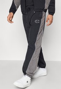 Man in zwarte en grijze trainingsbroeken met wit logo en witte sneakers wandelt over een lichtgrijze vloer tegen een effen achtergrond.