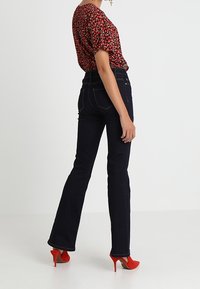 Mörkblå utsvängda jeans med hög midja, kompletterat med en kort blus i röd blommönster och ljusgröna högklackade skor.