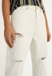 Bílé džínové kalhoty z denimu s opranými fleky na obou kolenou. Materiál je texturovaný a silný. Kalhoty mají standardní kapsy a zapínání na knoflík.