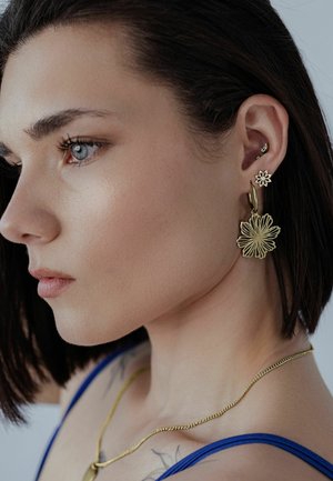 Frau mit kurz geschnittenem dunklem Haar, die goldene, blumenförmige Ohrringe, eine goldene Kettenhalskette und ein blaues Trägertop trägt, schaut nach links.