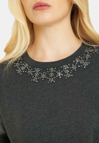 Ciemnoszary sweter z okrągłym dekoltem ozdobionym srebrnymi koralikami ułożonymi w dekoracyjny wzór wokół kołnierza.