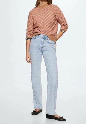 Femme portant un pull rayé rose, un jean taille haute bleu clair et des ballerines noires, debout avec une main sur la hanche.