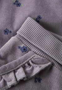 Lilla stoff med små mørkeblå blomstertrykk. Tekstilet har en ribbet, teksturert midje med en rysjekant.