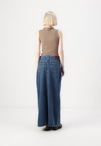Rjav brezrokavnik v kombinaciji z visokim pasom in širokimi modrimi jeans hlačami iz denima, ki imajo dva zadnja žepa in usnjen znak blagovne znamke.