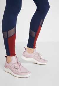 Nohy v námořnických modrých a červených legínách s síťovými panely a světle růžovými sportovními teniskami s bílou podrážkou na jednobarevném šedém podlaze.