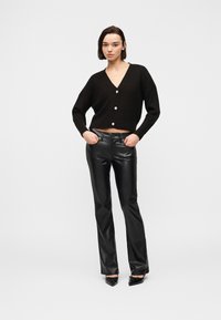 Γυναίκα που στέκεται με τα χέρια στις τσέπες, φορώντας μαύρη κοντή ζακέτα, μαύρο δερμάτινο παντελόνι και μαύρες μυτερές γόβες σε λευκό φόντο.