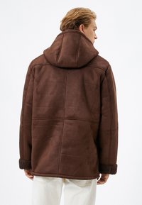 Brun faux suede jakke med hætte, der har sømdetaljer og fluffy manchetter. Teksturen virker blød og varm.