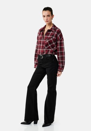 Frau trägt ein rot-schwarz kariertes Hemd mit Knöpfen, schwarze ausgestellte Hosen und schwarze spitze Stiefel und steht vor einem weißen Hintergrund.