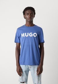 Kék pamut póló, elöl egy nagy fehér "HUGO" logóval. Kerek nyakú kialakítás, rövid ujjakkal és laza szabással.